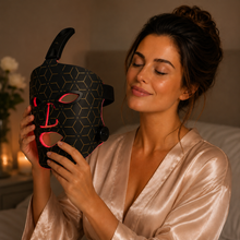 A woman in a satin robe holds a black LED light therapy face mask with gold geometric patterns and pink glowing light accents in a warm, dimly lit room.