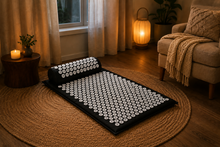 A black acupressure mat with white circular massage nodes is shown in a cozy, warmly lit living room setting on a woven jute rug, next to an armchair and a lit lantern lamp.