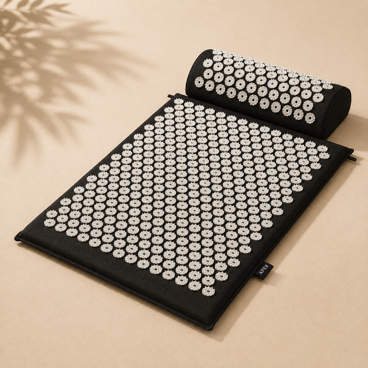 A black acupressure mat with a matching cylindrical pillow, both covered in white circular massage nodes, displayed on a beige background with soft leaf shadows.