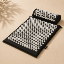 A black acupressure mat with a matching cylindrical pillow, both covered in white circular massage nodes, displayed on a beige background with soft leaf shadows.