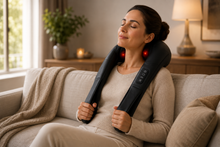 A woman sits on a beige sofa in a living room, using a black neck and shoulder massager with red illuminated nodes. The setting includes a window, a lamp, and a potted plant in the background.