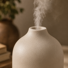 A close-up of a textured ceramic diffuser or vase with white mist rising from the top, set against a warm, softly blurred background with a plant and books.