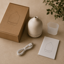 A minimalist essential oil diffuser set arranged on a warm beige surface, including a matte cream diffuser bottle, kraft paper box, white USB charging cable, small frosted cup, and printed instruction card. A leafy branch appears in the top-right corner.